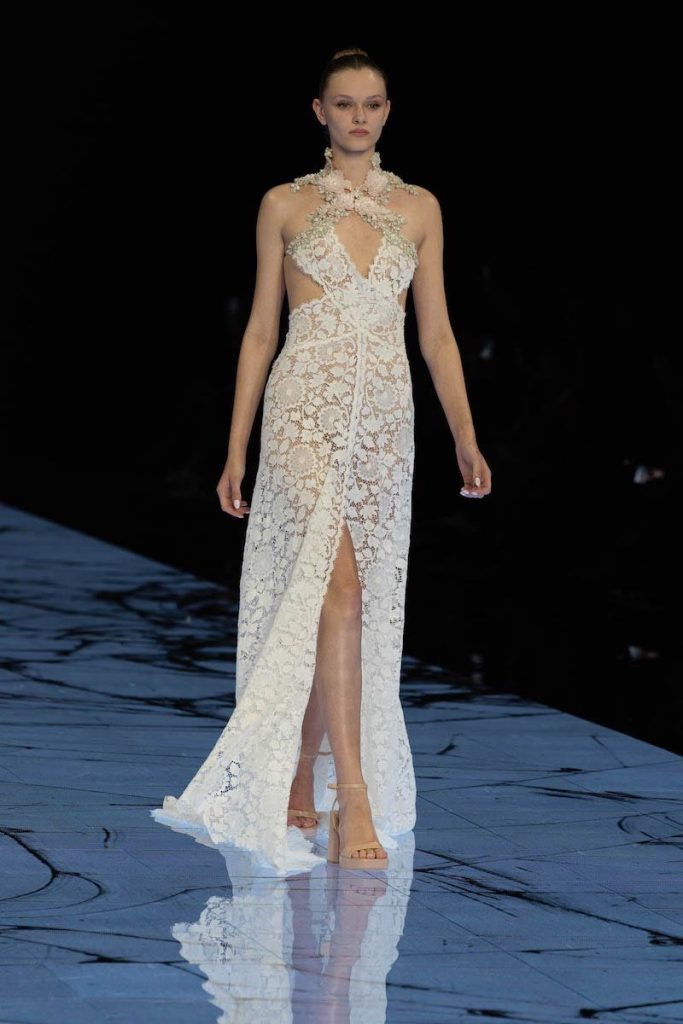 In questa foto una modella sfila indossando un abito da sposa Marco y Maria 2024 in pizzo floreale con dettaglio cut out sul busto ed elementi floreali lungo la scollatura ad incrocio sul collo
