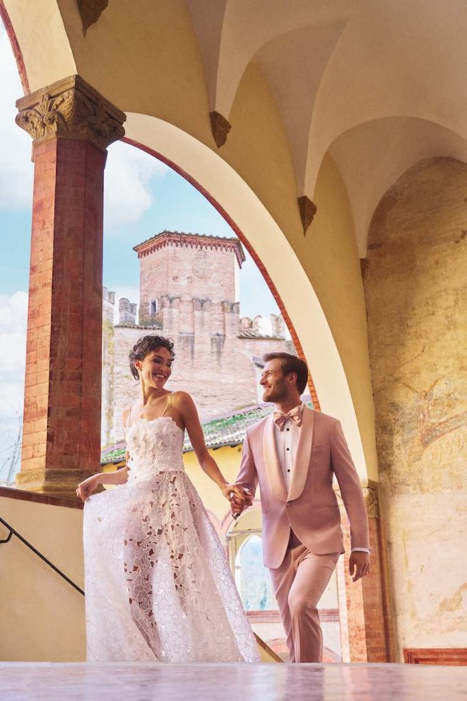 In questa foto una coppia di sposi si sale felice le scale di un antico palazzo. Lei indossa un abito scivolato in pizzo Sangallo; lui uno smoking di colore rosa