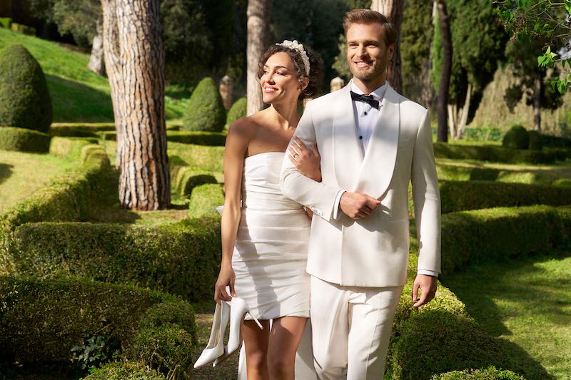 In questa foto due sposi camminano a braccetto nel parco di una villa. Lei indossa un abito corto drappeggiato e tiene le sue décolleté nella mano destra. Lui indossa uno smoking di colore bianco con revers sciallati