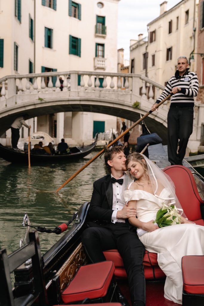 In questa foto due sposi sorridenti su una gondola a Canal Grande con alle spalle un gondoliere vestito a tema: lo scatto è stato realizzato da Sara Sganga, tra i migliori fotografi di matrimonio a Venezia