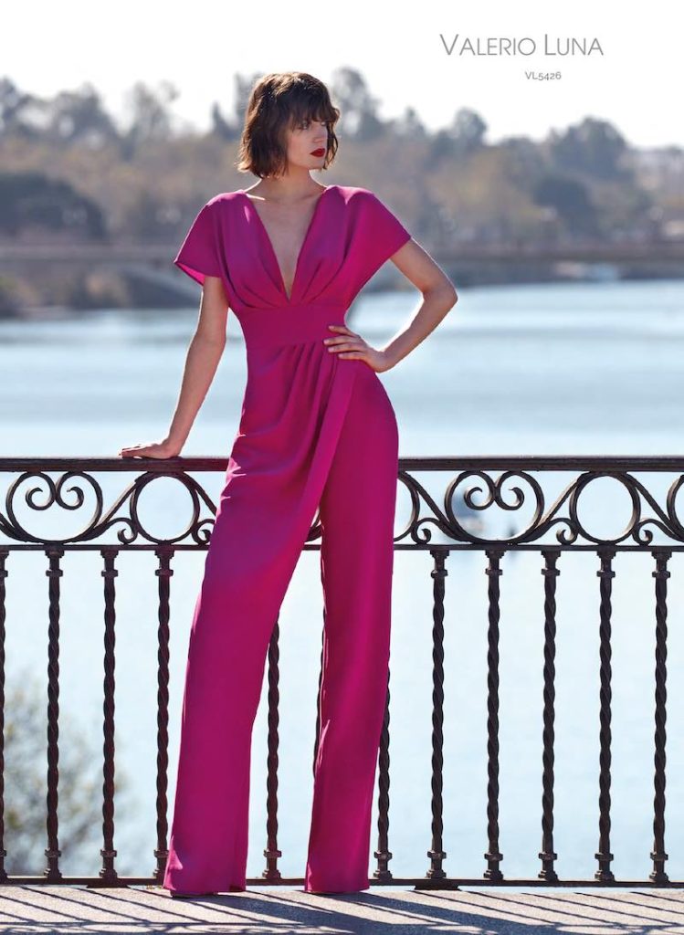 In questa foto una modella posa indossando una tuta di Valerio Luna con maniche corte, scollo a V e drappeggio frontale di colore fucsia per un look Barbiecore da cerimonia