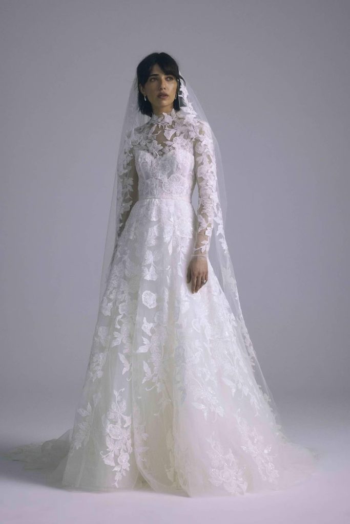 In questa foto una modella posa indossando un abito da sposa di Amsale ad A con maniche e tulle ricamato in pizzo abbinato ad un velo con bordo in pizzo