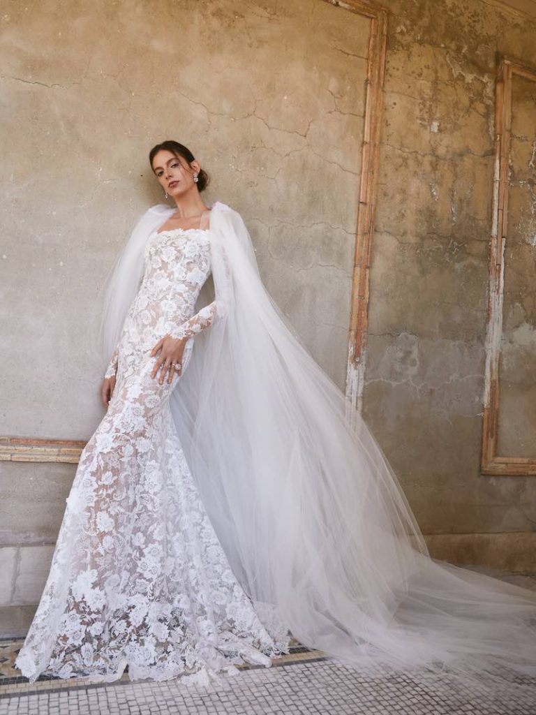 In questa foto una modella posa indossando un abito da sposa a sirena in pizzo floreale di Oscar de la Renta con maniche e mantella di tulle