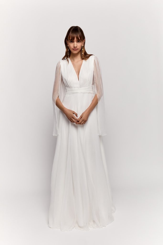 In questa foto una modella posa indossando un abito da sposa scivolato in chiffon con maniche trasparenti e scollo a V su corpetto drappeggiato