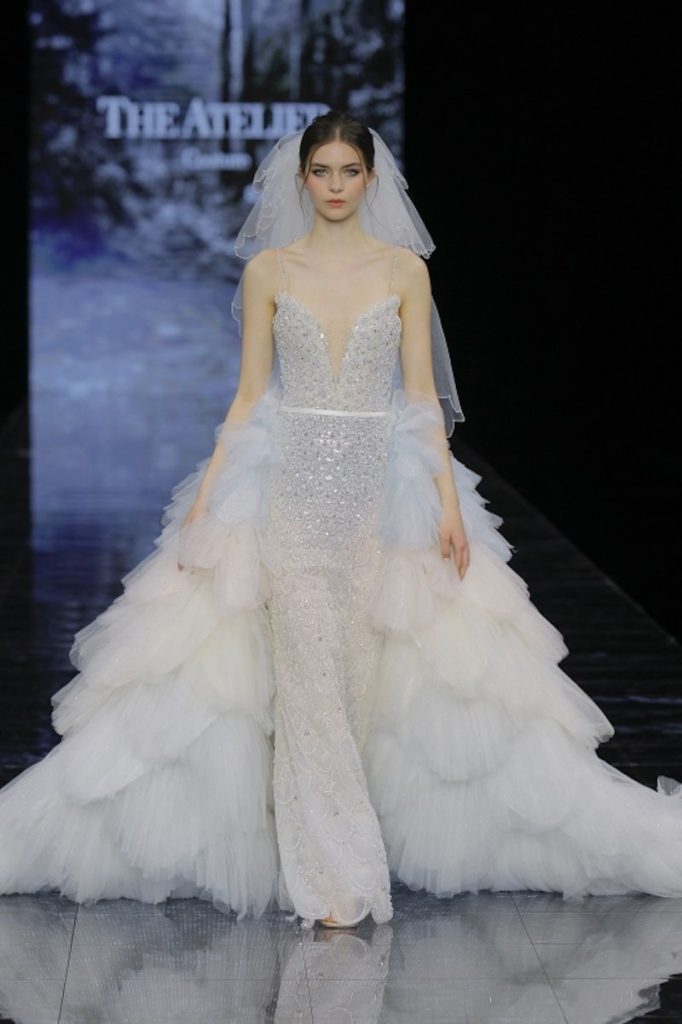 In questa foto una modella sfila indossando un abito da sposa a sirena Atelier Couture con perline e paillettes di colore celeste e sopragonna di tulle a balze abbinato