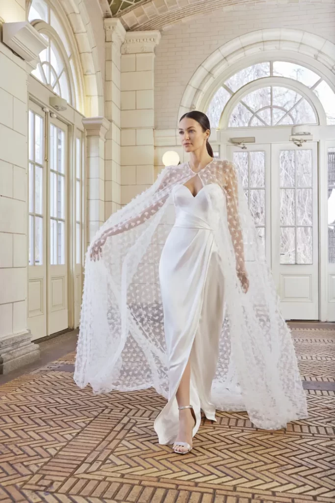 In questa foto una modella posa indossando un abito da sposa scivolato in seta di Sareh Nouri con scollo a cuore abbinato ad una mantella in tulle con ricami floreali all over