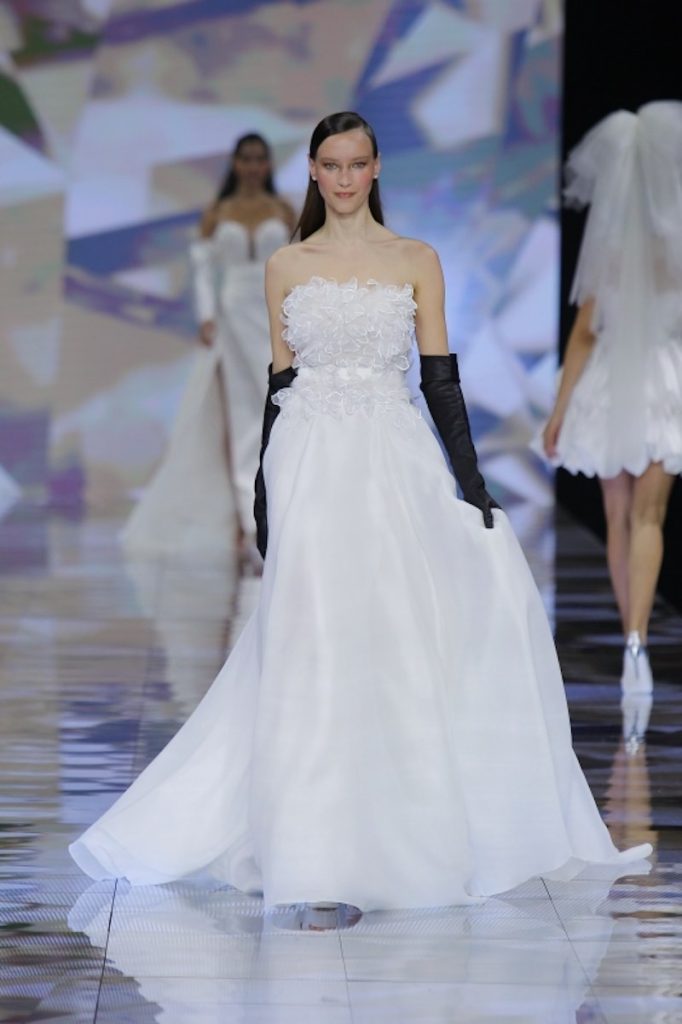 In questa foto una modella indossa un abito da sposa a trapezio di Nicole Milano con gonna in organza e corpetto in pizzo 3D abbinato ad un paio di guanti neri 