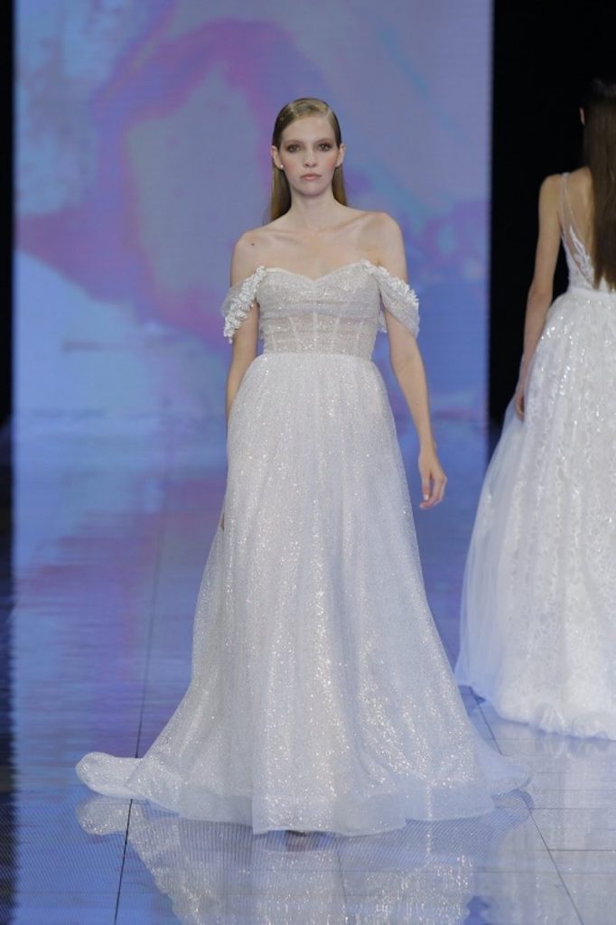 In questa foto una modella sfila indossando un abito da sposa con tessuto glitter di Nicole Milano con scollo off shoulder