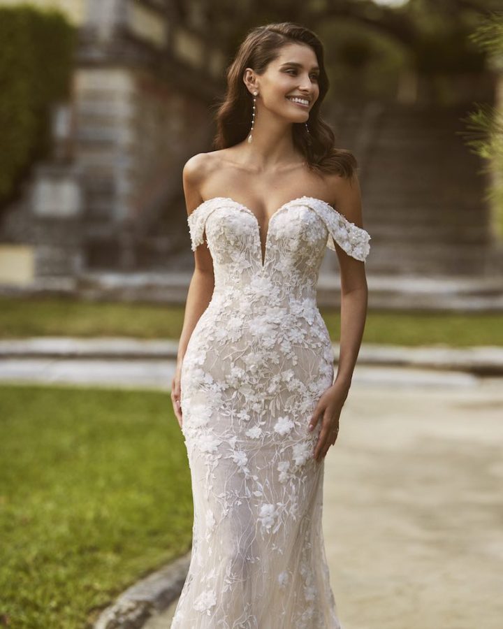 In questa foto una modella posa indossando un abito da sposa a sirena Aire Barcelona con scollo off shoulder ed elementi floreali 3D