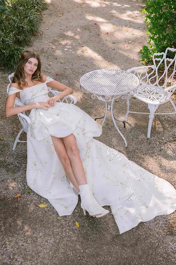 In questa foto una modella posa seduta su una sedia in ferro battuto in un giardino indossando un abito da sposa mullet di Monique Lhuillier con microfiori dipinti