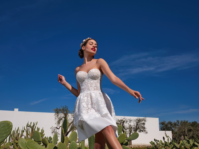 In questa foto una modella posa indossando un abito da sposa di Mysecret Sposa modello corto con scollo a cuore senza spalline e in pizzo floreale