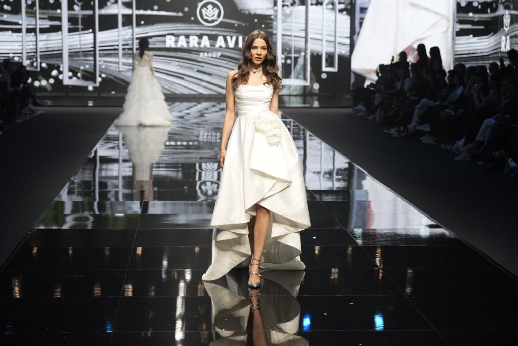 In questa foto una modella sfila indossando un abito da sposa mullet di Rara Avis con corpetto drappeggiato e maxi applicazione sul fianco sinistro