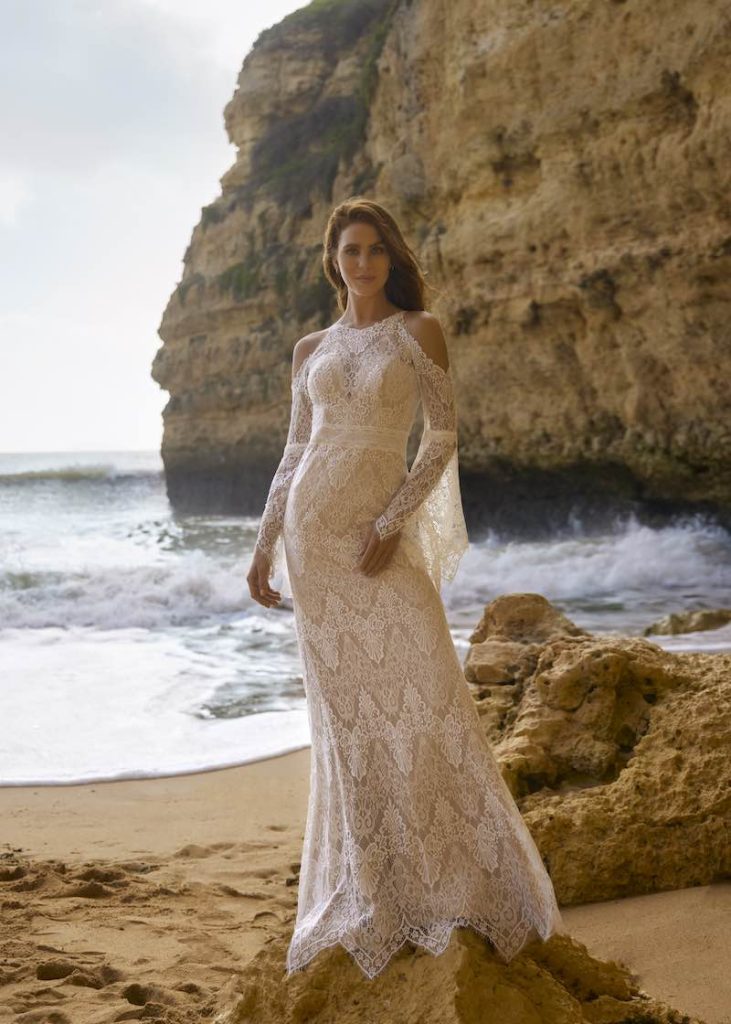 In questa foto una modella posa sulla spiaggia indossando un abito da sposa a sirena in pizzo di Cherish Love Collection by GBS Herve Paris con ma niche a campana