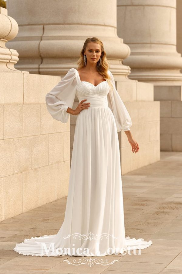 In questa foto una modella posa con un abito da sposa di Monica Loretti modello scivolato in chiffon con maniche lunghe a sbuffo e scollo a cuore