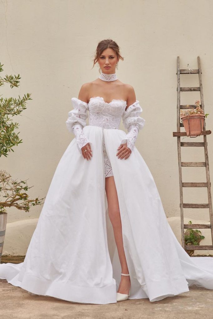 In questa foto una modella posa indossando un abito da sposa di Claudio Di Mari in pizzo Sangallo con maniche staccabili abbinate e sopragonna in taffetá