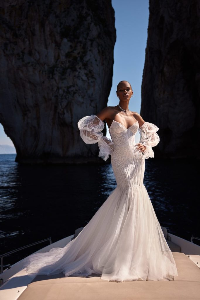 In questa foto una modella posa su una barca indossando un abito da sposa di Julie Vino a mezza sirena in tulle con ricami sparkling e maniche balloon rimovibili
