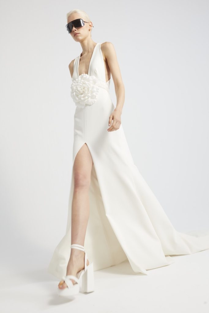 In questa foto una modella posa indossando un abito da sposa di Yolancris con spacco frontale, scollo a V e maxi applicazione a forma di camelia sul punto vita