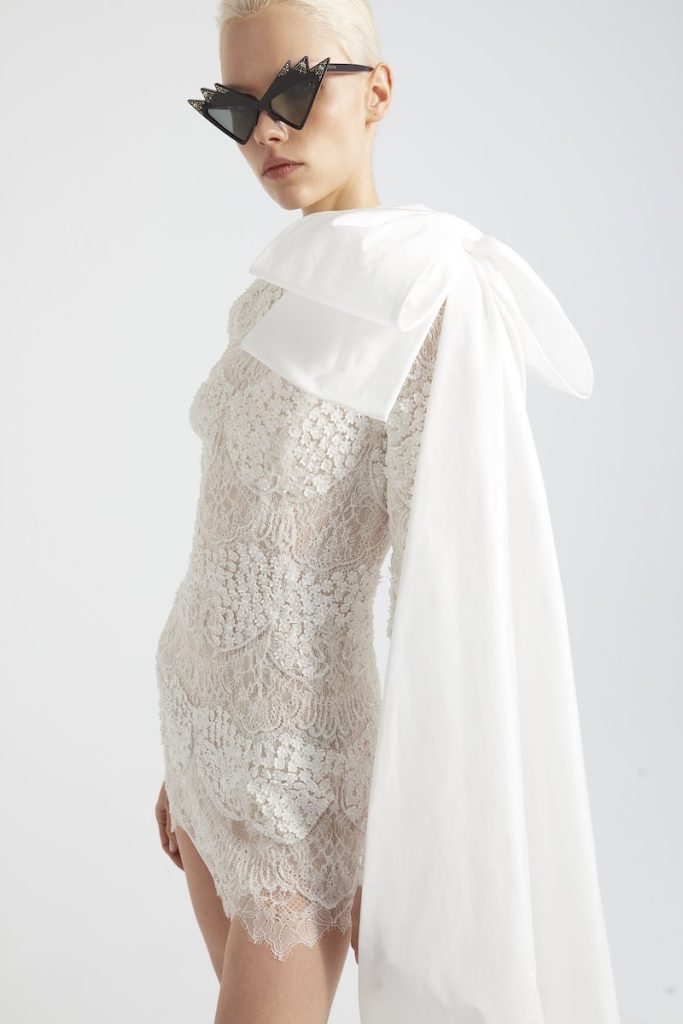 In questa foto una modella posa indossando un abito da sposa corto in pizzo francese di Yolancris con maxi fiocco sulla spalla sinistra
