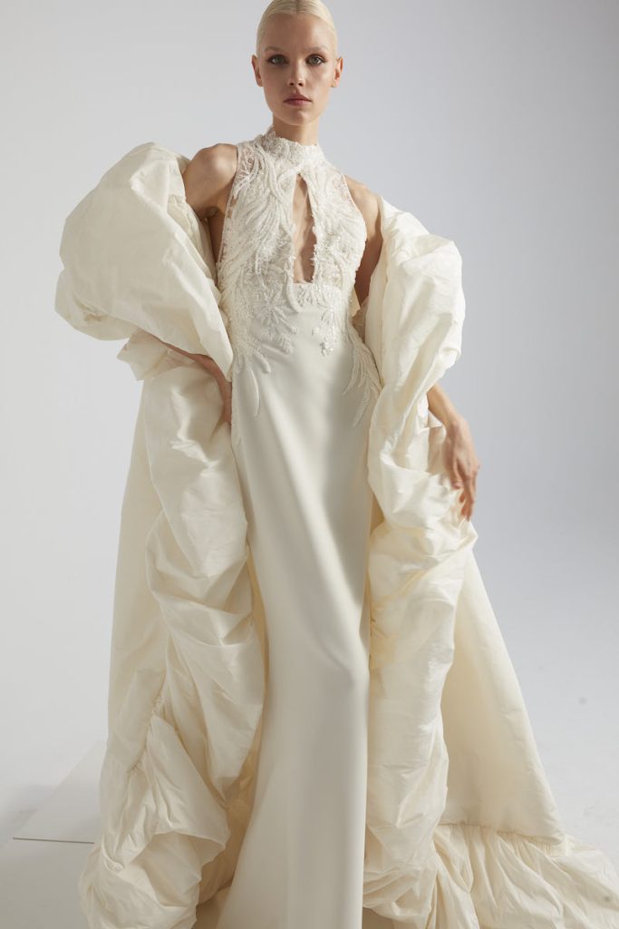 In questa foto una modella posa indossando un abito a sirena di Yolancris con busto ricamato in pizzo, scollo all'americana abbinato ad un cappotto da sposa over
