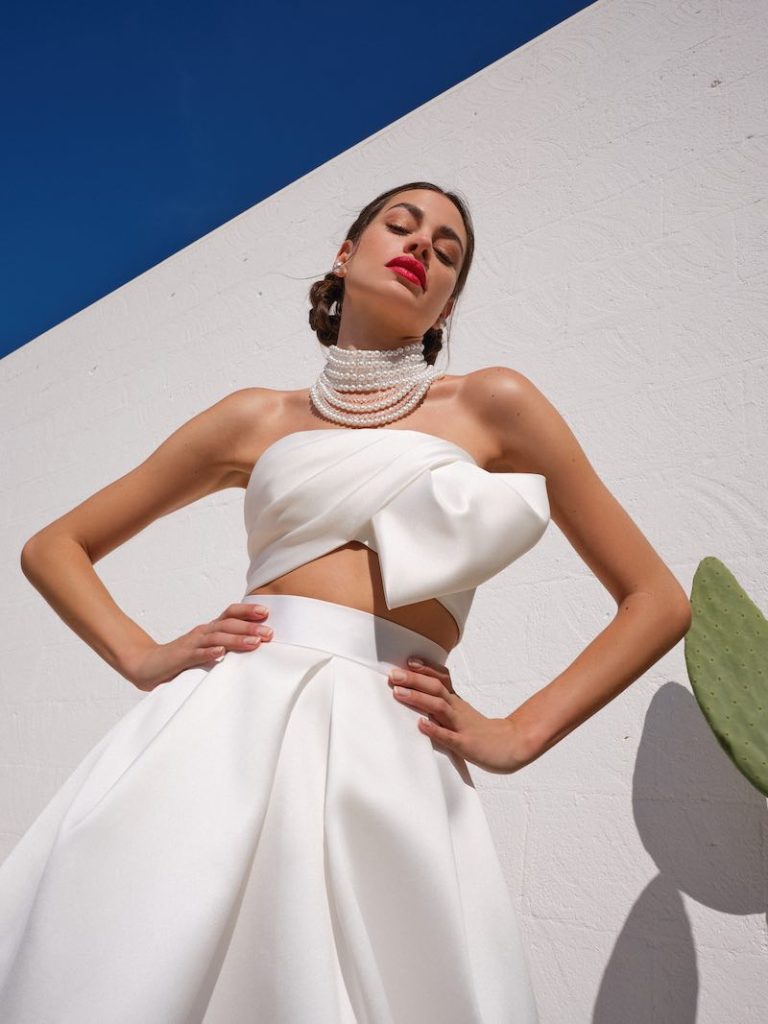In questa foto una modella posa indossando un abito da sposa due pezzi con crop top di Mysecret Sposa. Il top presenta un grande fiocco destrutturato