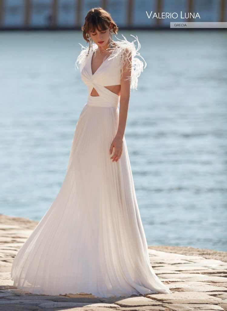 In questa foto una modella posa indossando un abito da sposa scivolato di Valerio Luna con effetto cut out sul punto vita, gonna plissé ed effetto plumage sulle spalle