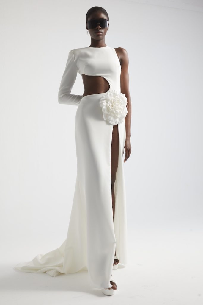 In questa foto una modella posa indossando un abito da sposa a sirena di Yolancris con effetto cut-out sul fianco destro, maniche lunga sul braccio destro e maxi camelia sul fianco sinistra