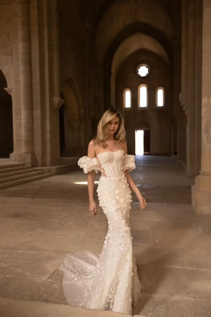 In questa foto una modella posa indossando un abito da sposa di Eva Lendel a mezza sirena con bustier, maniche balloon ed elementi 3D all over