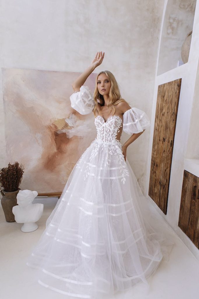 In questa foto una modella posa indossando un abito da sposa Agnieszka Swiatly con corpetto con foglie e petali 3D, gonna in organza a righe e maniche balloon removibili