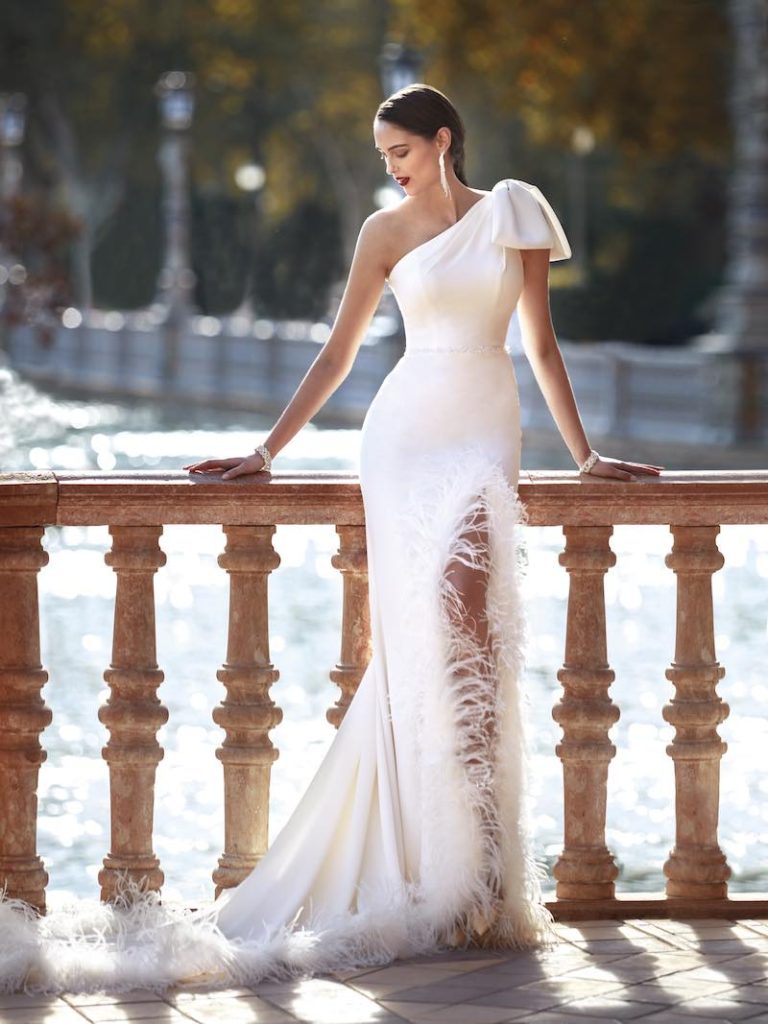 In questa foto una modella posa indossando un abito da sposa a sirena di Manu Garçia con maxi fiocco sulla spalla, spacco frontale e plumage lungo lo spacco