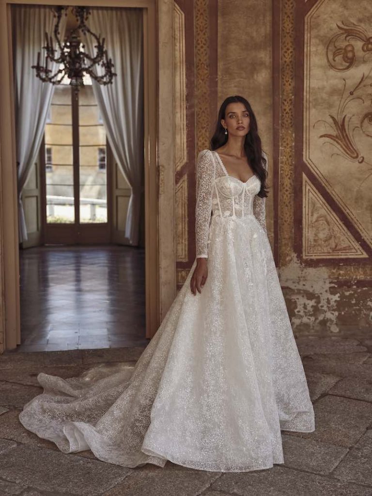 In questa foto una modella indossa un  abito da sposa ad A con tessuto sparkling, bustier, scollo a cuore e maniche lunghe di Dominiss