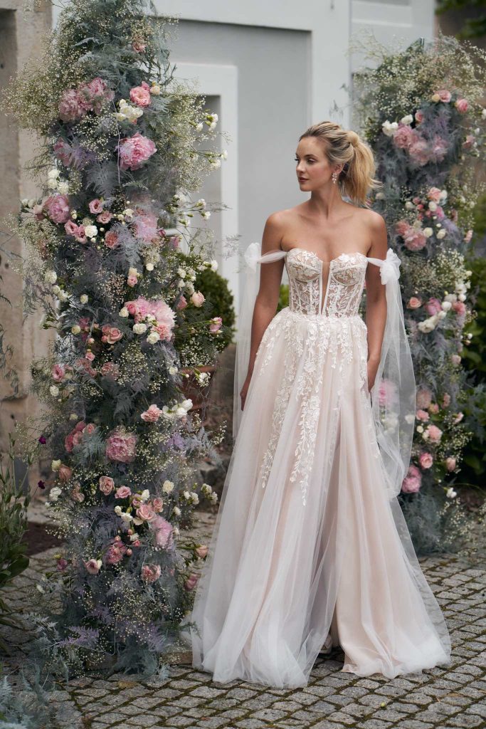 In questa immagine un abito da sposa con corpetto interamente ricamato in pizzo con decori floreali e delle alette in tulle che partono dalle braccia. 