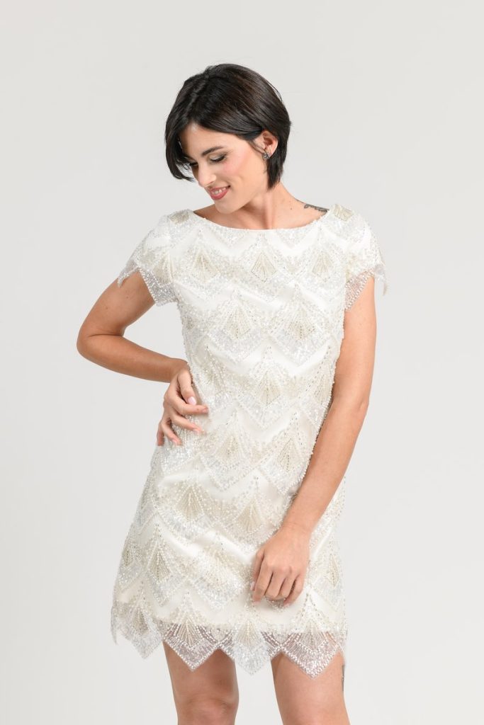 In questa foto una modella posa indossando un abito da sposa modello tubino della collezione Unique di Benedetta Passalacqua. L'abito è corto sopra al ginocchio ed è ricamato con perline all over