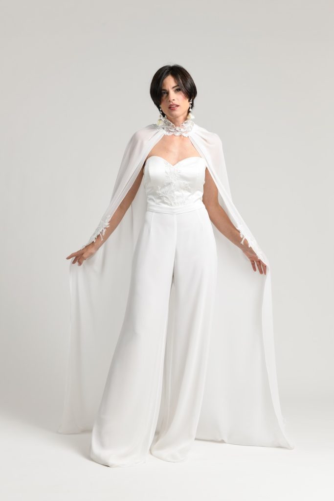 In questa foto una modella posa indossando una tuta da sposa della collezione Unique dell'Atelier Benedetta con corpetto in mikado ricamato con scollatura a cuore e pantaloni in chiffon. La modella indossa anche una cappa in chiffon con collarino con fiore 3D