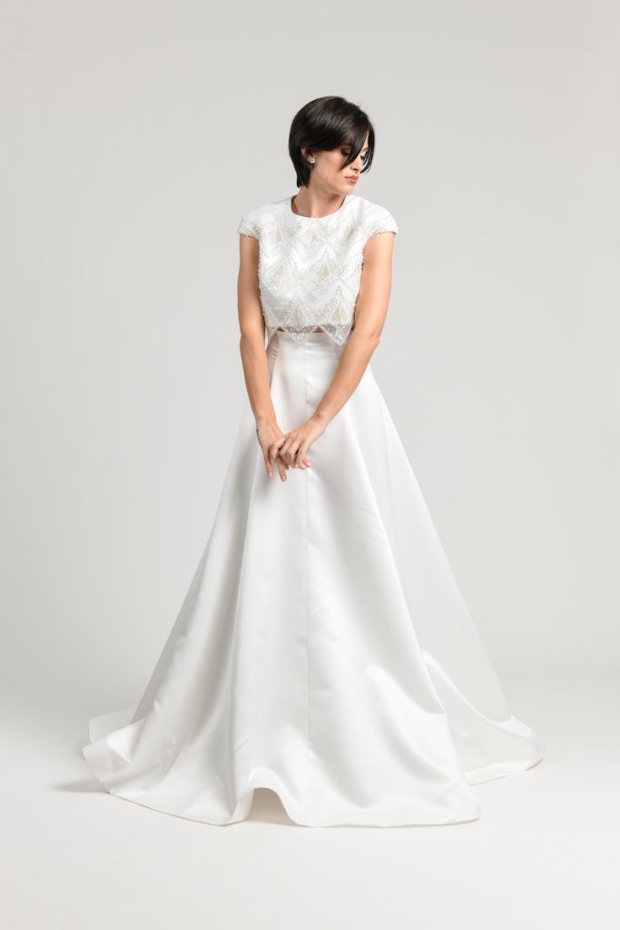 In questa foto una modella indossa un abito da sposa due pezzi con crop top a maniche corte con ricamo di perline e una gonna in mikado dell'Atelier Benedetta