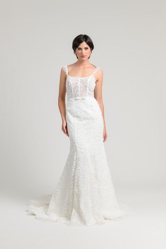In questa foto una modella posa indossando un abito da sposa a semi sirena in tulle ricamato con scollo quadrato e micro fiocco in vita