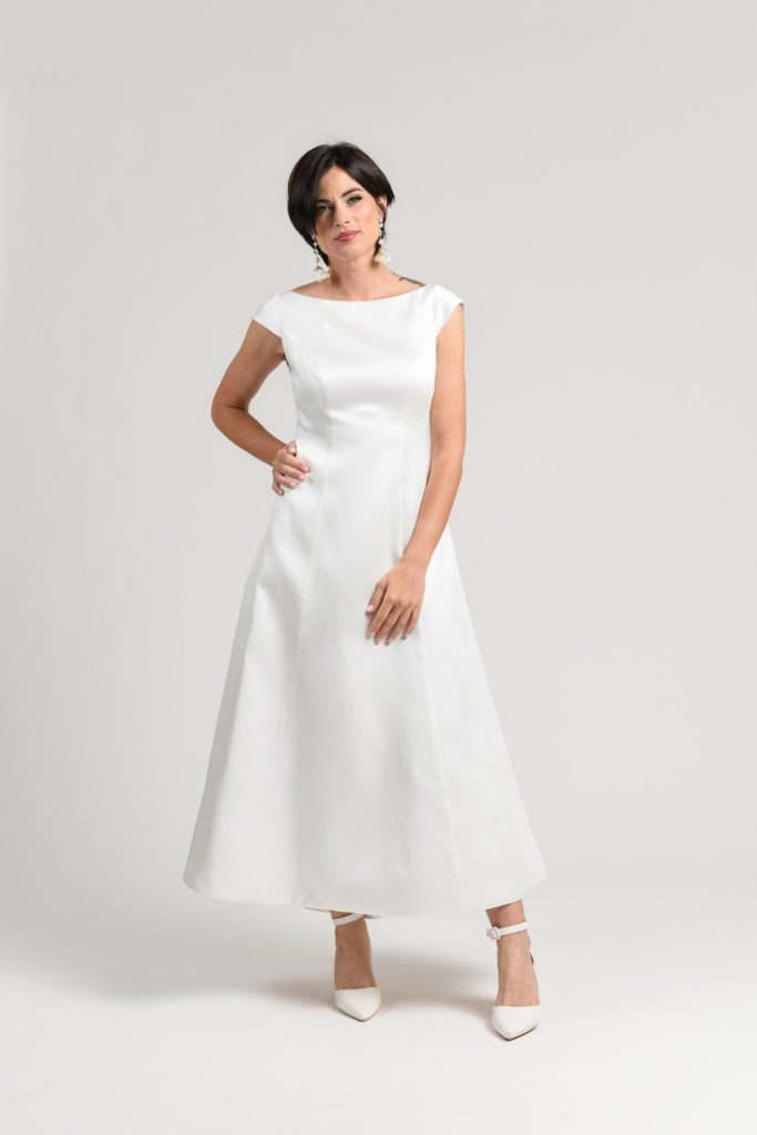 In questa foto una modella posa indossando un abito da sposa midi alla caviglia della collezione Unique dell'Atelier Benedetta. L'abito presenta una scollatura a barchetta e una gonna svasata