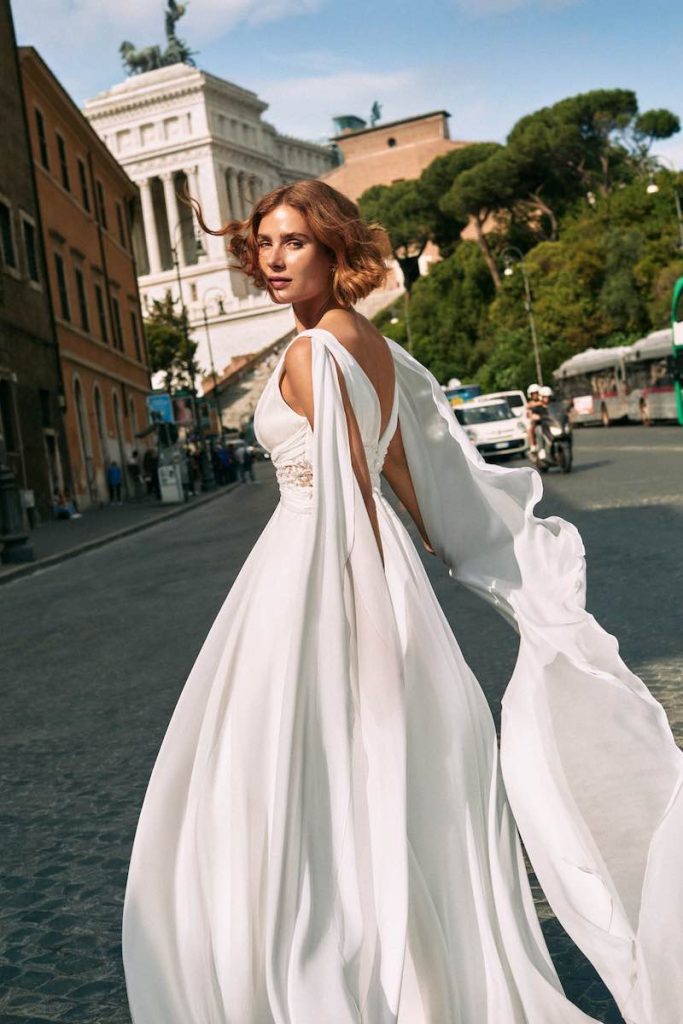 In questa foto una modella posa correndo per le strade di Roma sullo sfondo dell'Altare della Patria indossando un abito da sposa Atelier Emé in chiffon con scollatura e a cuore e morbide ali