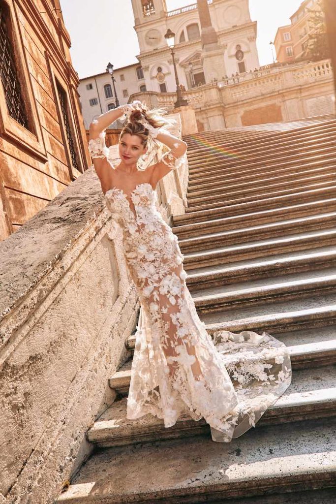 In questa foto una modella posa sulle scale di Piazza di Spagna indossando un abito della nuova collezione sposa Atelier Emé 2024 modello a sirena con maniche a guanto staccabili, in tulle con applicazioni di fiori 3D