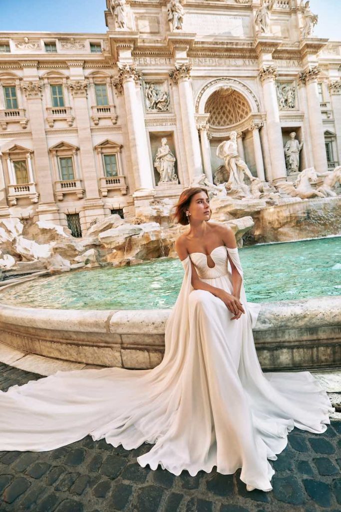 In questa foto una modella posa seduta sul bordo di Fontana di Trevi indossando un abito da sposa Atelier Emé 2024 in chiffon con scollatura e a cuore e morbide ali
