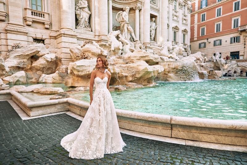 In questa foto una modella posa davanti alla Fontana di Trevi indossando un abito nuova collezione sposa Atelier Emé 2024 dalla linea ad A e interamente ricamato in pizzo