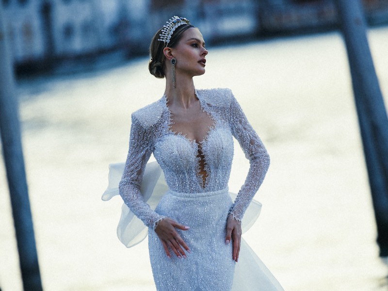 In questa foto una modella posa a Venezia indossando un abito da sposa Maison Signore 2024 con maniche e interamente ricamato di perline. La modella indossa anche una tiara di pietre