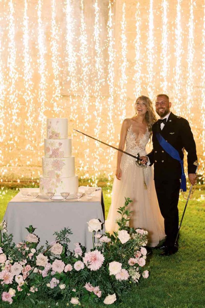 In questa foto una coppia di sposi al taglio della loro torta nuziale a quattro piani decorata con fiori 3D in crema di burro sullo sfondo di una cascata di luci e fuochi d'artificio