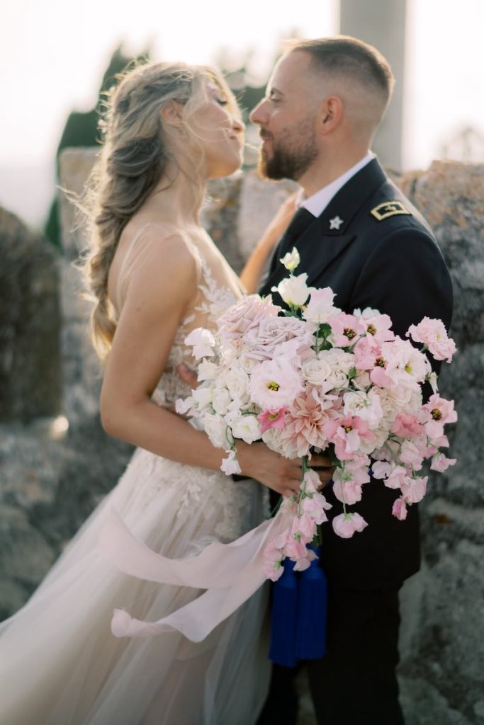 In questa foto due sposi si guardano e si abbracciano innamorati nel giorno del loro matrimonio. In primo piano è mostrato il suo bouquet con fiori nei toni del rosa stretti da un nastro di lino abbinato 