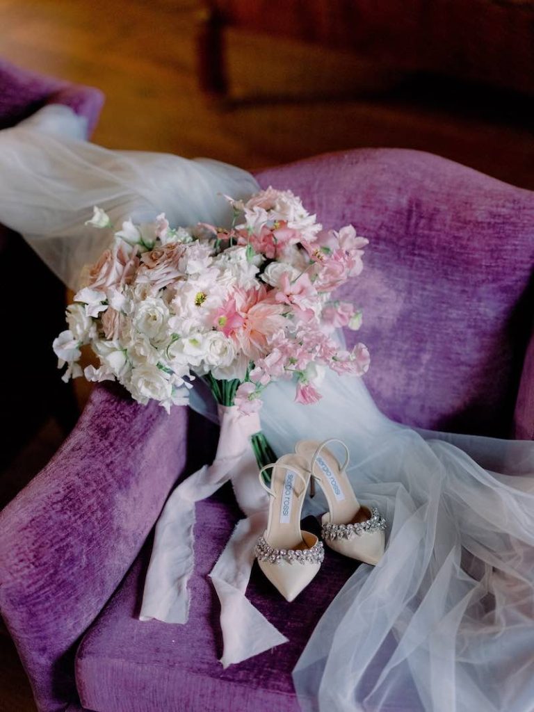 In questa foto un bouquet da sposa realizzato con fiori di colore rosa e bianco chiusi da un nastro di colore rosa e posizionato su una poltrona di colore viola al fianco delle scarpe gioiello della sposa 