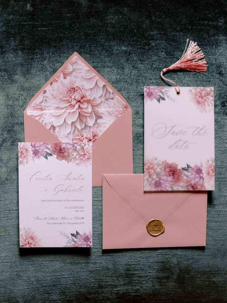 In questa foto una partecipazione di nozze di colore rosa decorata con fiori di dalie di colore rosa e la cui busta è chiusa da un bollino di ceralacca su cui è impresso il simbolo della Triforza di "The Legend of Zelda"