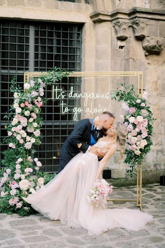 In questa foto due sposi fanno un casqué davanti al Photobooth realizzato con una scritta al neon che riporta "It's dangerous to go alone" decorato con fiori di colore rosa, bianco e lilla