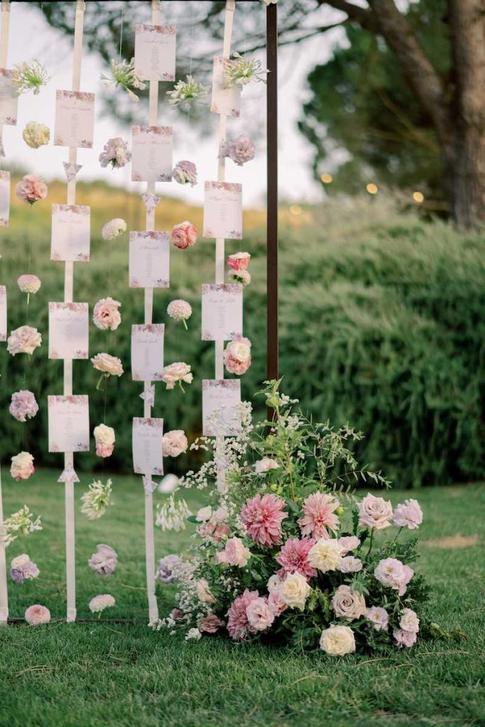 In questa foto un tableau de mariage in stile romantico decorato con fiori di colore rosa e lilla e composto da card su nastri che scendono da una struttura in ferro battuto