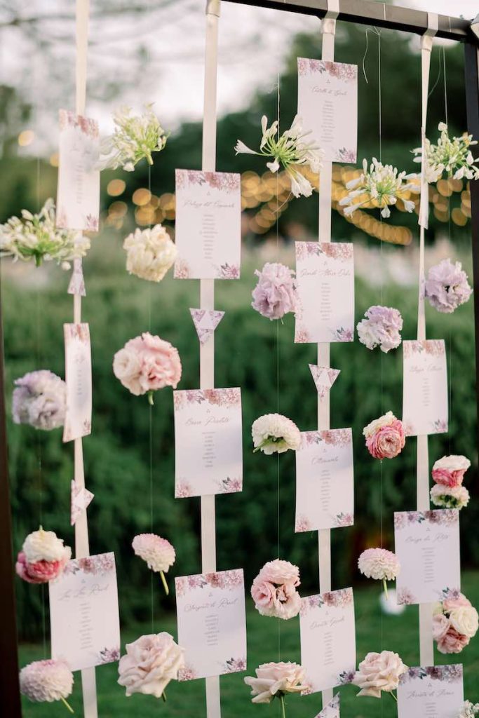 In questa foto il dettaglio di un tableau de mariage in stile romantico decorato con fiori di colore rosa e lilla e composto da card su nastri che scendono da una struttura in ferro battuto
