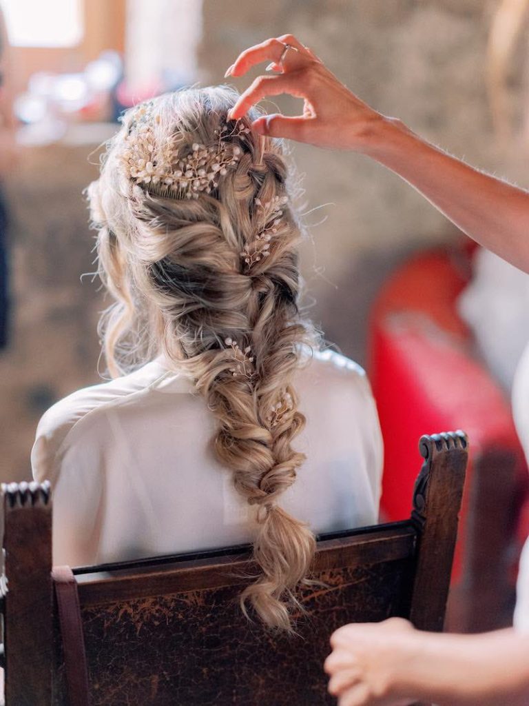 In questa foto una sposa di spalle durante la creazione della sua acconciatura: una treccia scomposta decorata con un accessorio di perline e cristalli