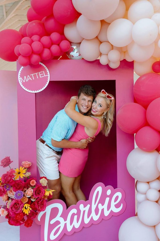 In questa foto una coppia di sposi in un Photobooth a forma di scatola di Barbie di colore fucsia e decorato con palloncini di abbinati 